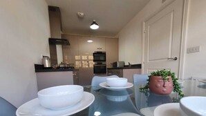 A modern kitchen with stainless steel appliances, a glass dining table with white plates and bowls, and a potted plant on the table. - Tranquil Oasis Modern and Spacious Retreat 4-Bedroom with Private Parking (Luton)