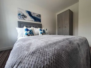 Una cama bien hecha con cubrecama gris y almohadas florales azules, cabecero gris, armario grande de color gris y un cuadro abstracto colgado en la pared.