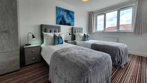 A hotel room with two beds, a grey wardrobe, a painting on the wall, and a window with a view of buildings. - Tranquil Oasis Modern and Spacious Retreat 4-Bedroom with Private Parking (Luton)