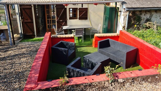 A covered patio area with a table and chairs, surrounded by a red border and a small garden.