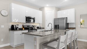 A modern kitchen with white cabinetry, a central island with bar stools, stainless steel appliances, and a clock on the wall. - 4 BD/ 4 BA/ Formosa Valley (7973 FV) (Kissimmee)