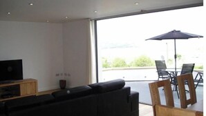 Ein modernes Wohnzimmer mit einer schwarzen Couch, einem Holztisch und einem Fernseher auf einer Holzkommode. Ein großes Fenster bietet Blick auf grüne Natur.