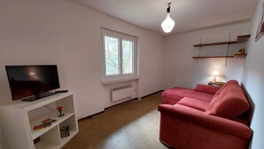Ein Wohnzimmer mit Fernseher auf einem weißen Schrank, einem rosafarbenen Sofa, einem Fenster mit Jalousien, einem Heizkörper und einer Hängeleuchte.
