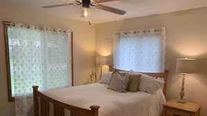 Une chambre à coucher avec un lit en bois, deux tables de chevet, un ventilateur de plafond et des rideaux aux fenêtres.