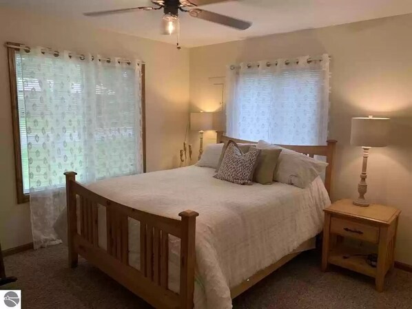 Un dormitorio con cama de madera, dos mesitas de noche, ventilador de techo y cortinas en las ventanas.