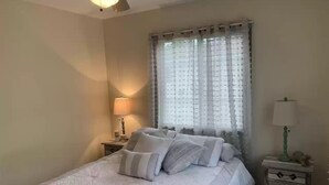 Une chambre à coucher comprenant un lit, deux tables de chevet, un ventilateur de plafond, une fenêtre avec des rideaux et une petite table de nuit avec une lampe.