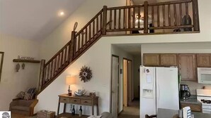 Un vaste espace de vie agrémenté d’un escalier en bois, une cuisine dotée d’appareils électroménagers blancs et un coin salon confortable avec une lampe et des objets décoratifs.