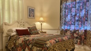 Un dormitorio con colcha estampada, un cabecero blanco ornamentado, una lámpara de noche y una cortina con motivos florales.