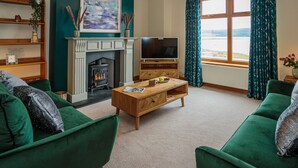 Una sala de estar con sofás verdes, una chimenea, un televisor y vista al mar desde una ventana.
