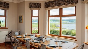Comedor con mesa de madera dispuesta para cuatro, ventanas amplias con vista a un lago y colinas, y un radiador en la pared.