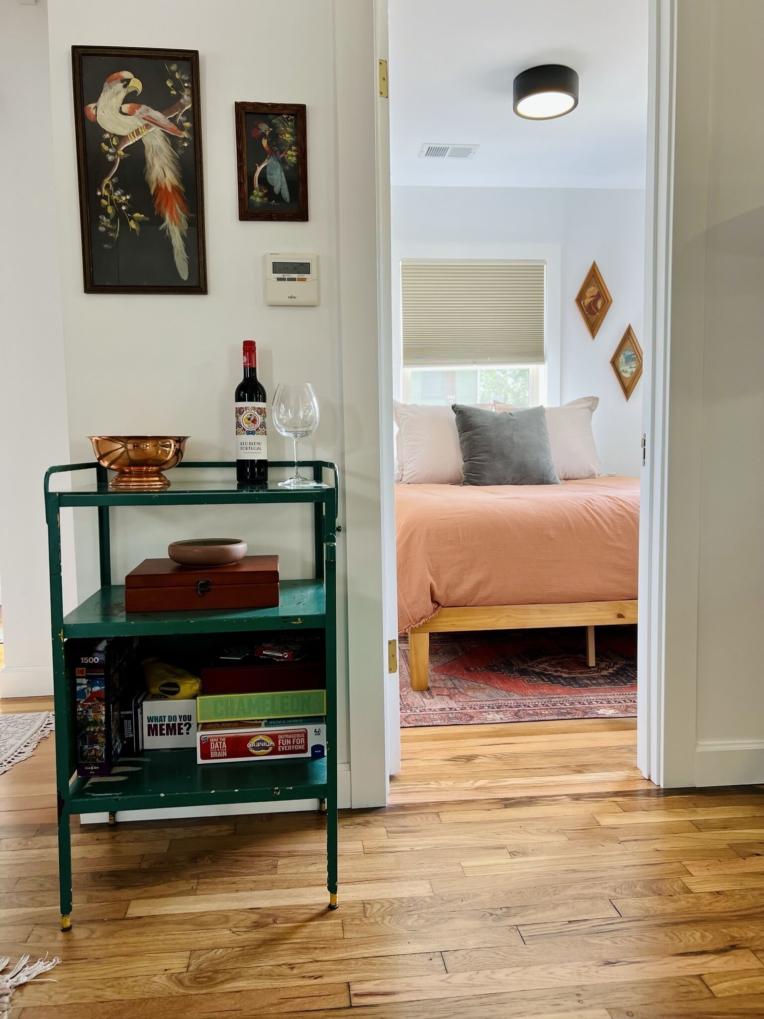 Une pièce avec un plancher en bois, un lit recouvert d’une couverture rose, une étagère en métal verte avec des livres et une bouteille de vin, et deux tableaux encadrés accrochés au mur.