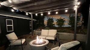 An outdoor patio area with a fire pit, two white cushioned chairs, and a covered seating area with string lights. - Renovated Retro Bungalow - Walk to Downtown! (Prescott)