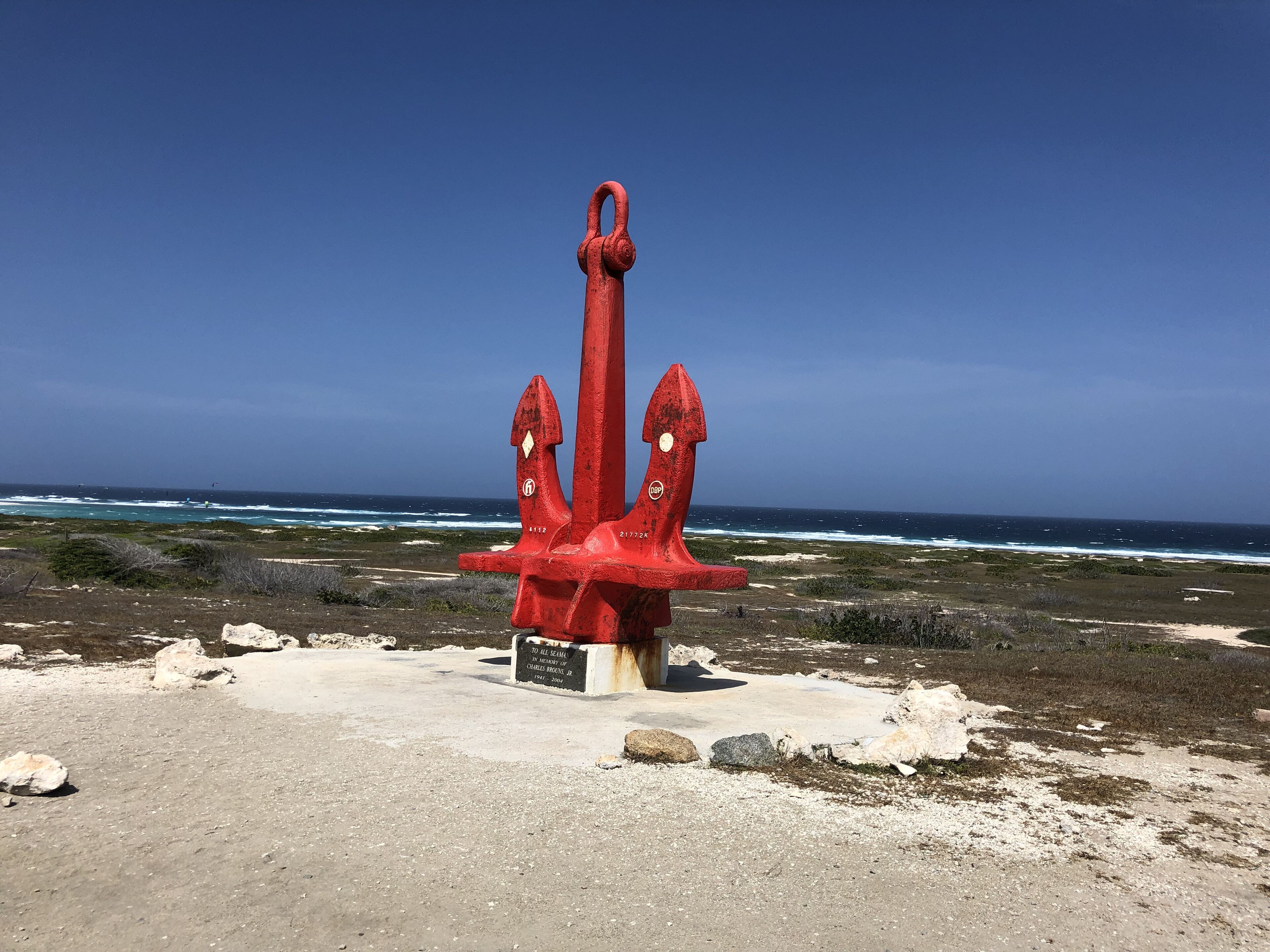 Een rood anker monument met inscripties, gelegen op een rotsachtig strand met de oceaan op de achtergrond.