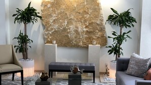 Een moderne woonkamer met een grote stenen muur, twee potplanten, een grijze bank, een glazen salontafel en een beige fauteuil.