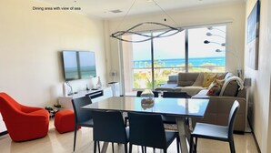 Een eethoek met uitzicht op zee, met een glazen tafel, zwarte stoelen, een rode stoel, een bank en een televisie aan de muur.