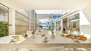 A sunlit outdoor dining area with a wooden table set for breakfast, overlooking a modern building and a view of the sea. - Seafront Villa Sunset Alcudia by Balearic Villas (Alcúdia)