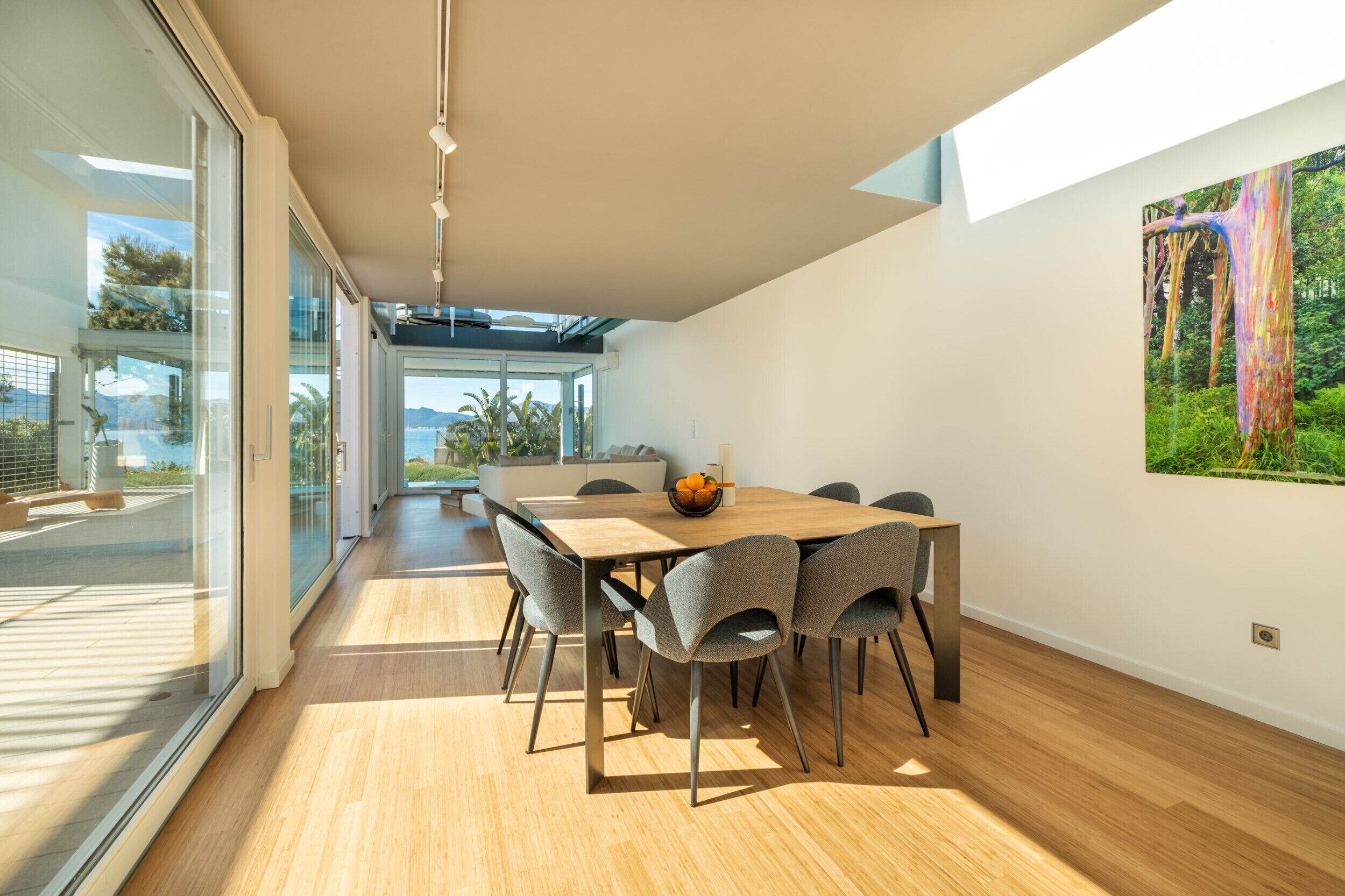 A modern dining area with a wooden table and grey chairs, large windows, and a view of the outdoors.