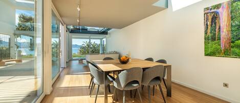 A modern dining area with a wooden table and grey chairs, large windows, and a view of the outdoors.