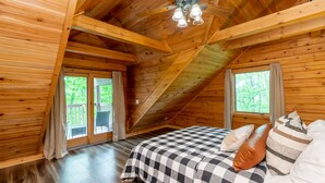 Un dormitorio rústico de cabaña de madera con colcha de cuadros, ventilador de techo y vista al exterior a través de una puerta corrediza de vidrio.