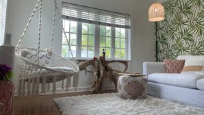 A modern living room with a hanging chair, a white sofa, and a large window with blinds. - Hot tub, tiki bar, outdoor shower, water access! (West Palm Beach)