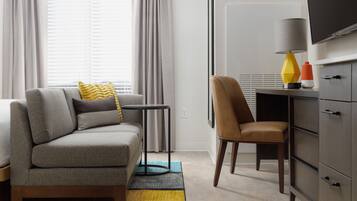 A modern hotel room with a grey sofa, a brown chair, a small table, a lamp, and a TV on a cabinet.