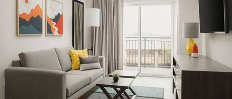 A modern hotel room with a grey sofa, a small coffee table, a TV, and a balcony with curtains.