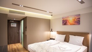 A hotel room with a wooden floor, a bed with a patterned bedspread, a nightstand with a lamp, and a painting of a sunset on the wall.