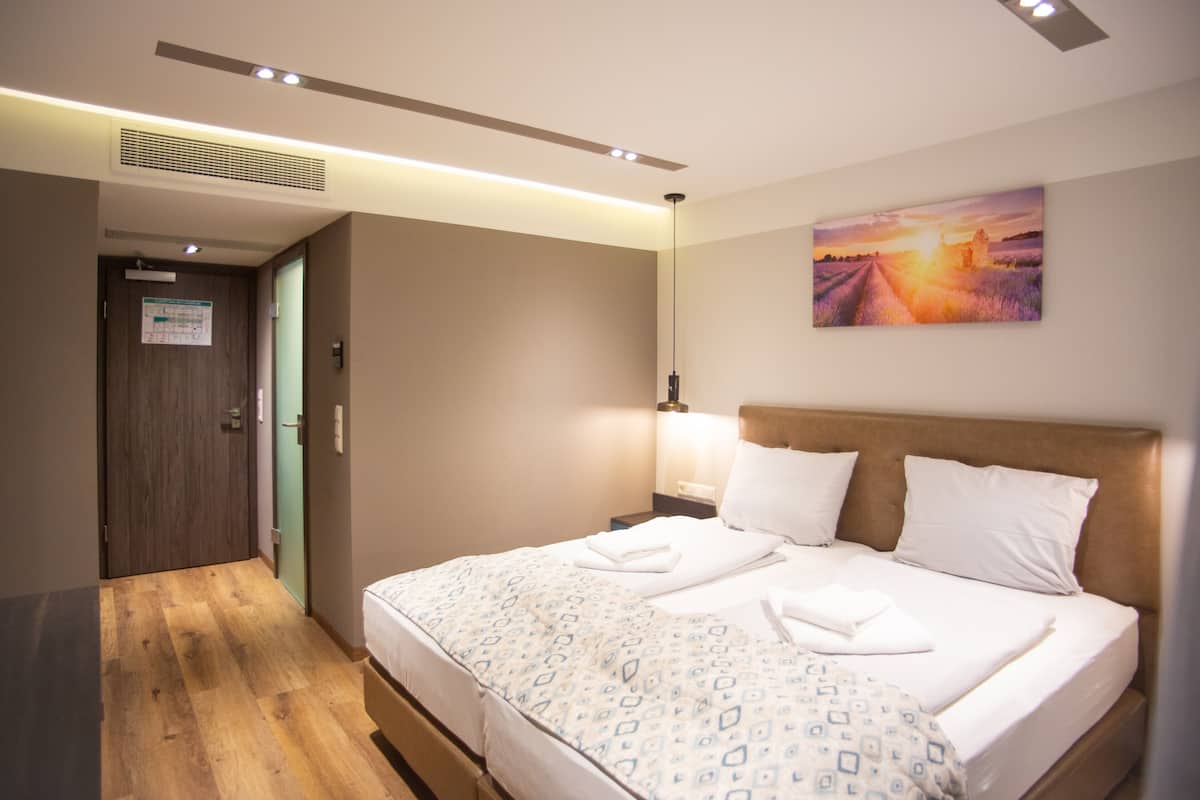 A hotel room with a wooden floor, a bed with a patterned bedspread, a nightstand with a lamp, and a painting of a sunset on the wall.