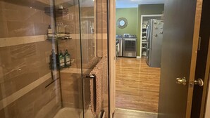 A hallway with a glass shower door, a wooden floor, and a kitchen visible through an open door. - Minutes from Churchill Downs & Downtown Lou! (Louisville)
