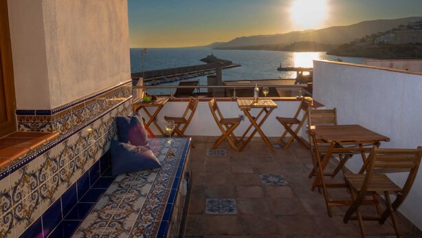 A terrace with a view of the sea at sunset, featuring a tiled wall, a wooden bench, and a table with chairs. - Peniscola Intramuros Suites (Peñíscola)