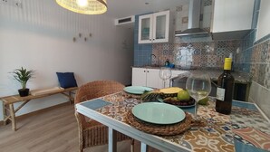 A compact kitchen with a dining area, featuring a wicker chair, a small table with a patterned tablecloth, and a hanging light fixture. - Peniscola Intramuros Suites (Peñíscola)