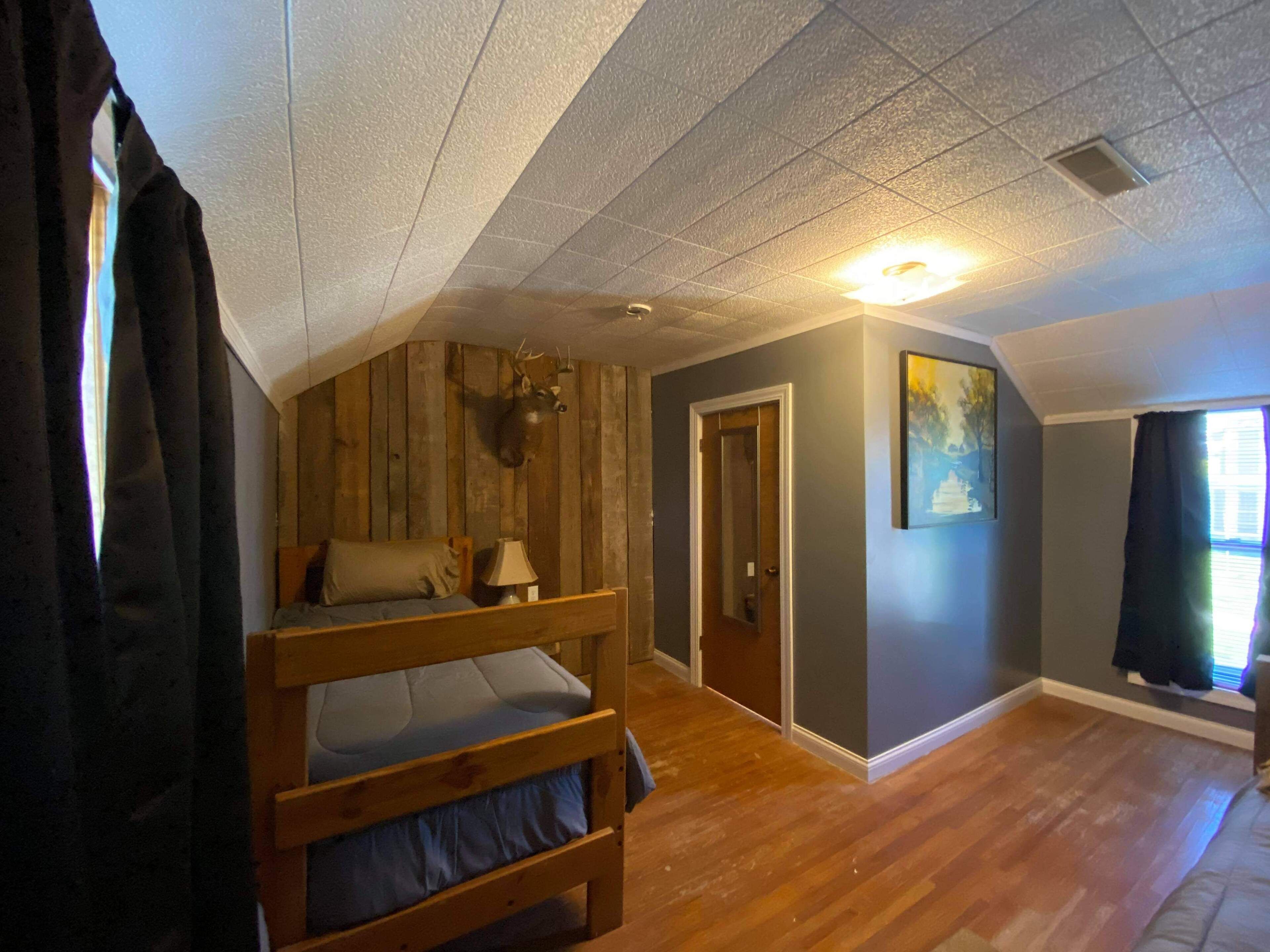 Quarto com beliche, paredes de madeira, cabeça de veado pendurada, porta e janela com cortinas.