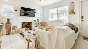 Une chambre moderne avec une cheminée, un lit recouvert d’une couverture blanche, un téléviseur fixé au mur et une grande fenêtre avec des stores.
