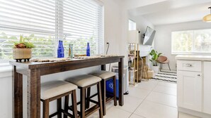 Une cuisine moderne avec une table en bois, des tabourets de bar et une grande fenêtre équipée de stores.