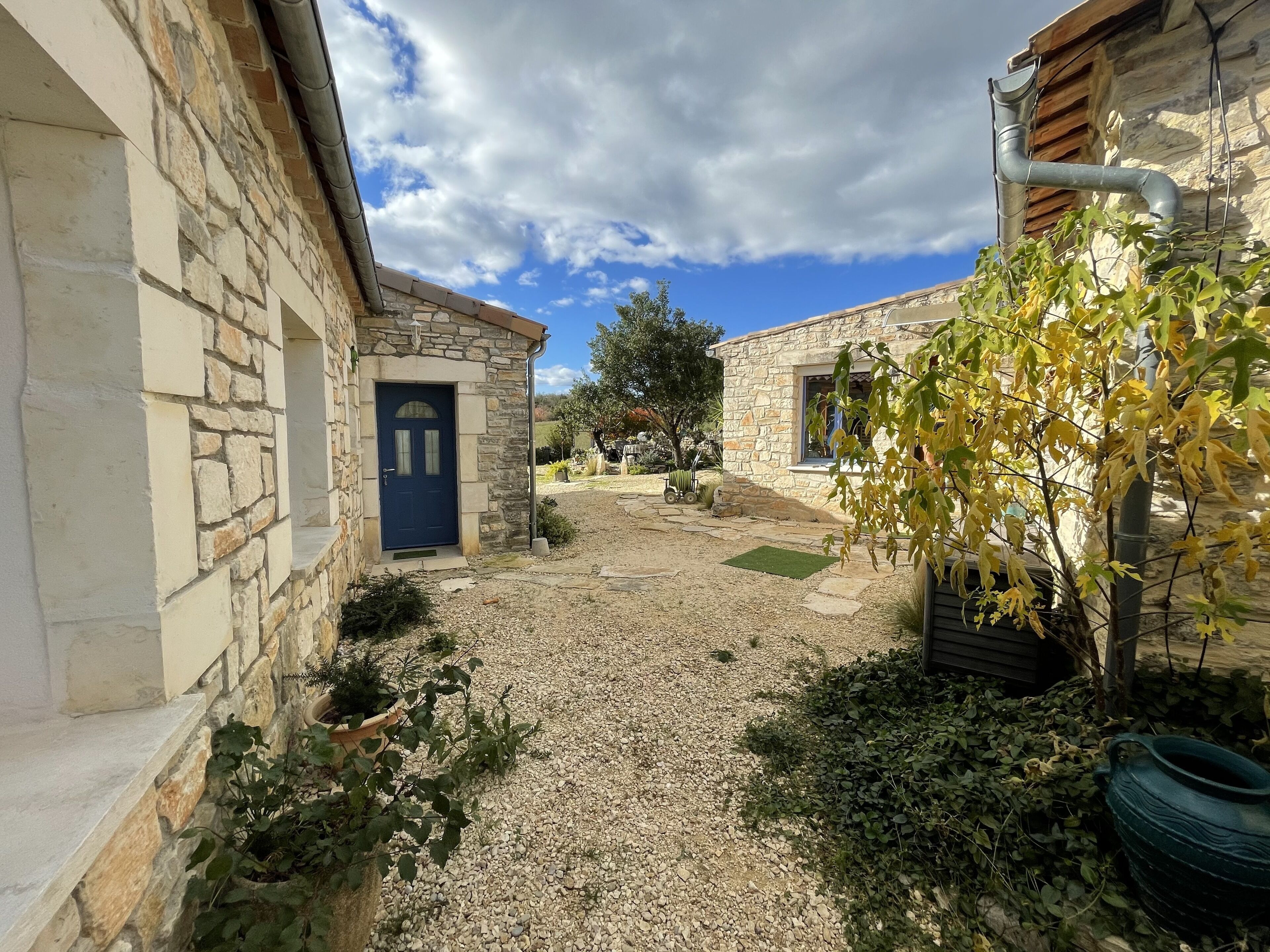 A stone building with a blue door, a gravel courtyard, and surrounding vegetation.