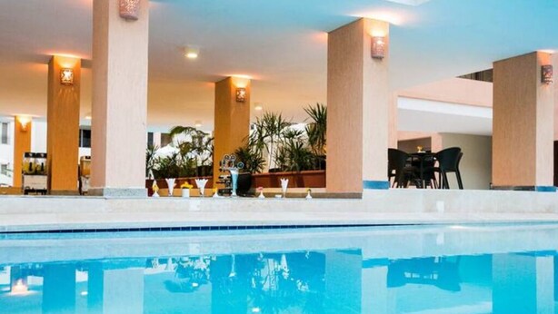 Zona de piscina de hotel con una piscina azul cristalina, rodeada de pilares color beige y una zona de estar con sillones.