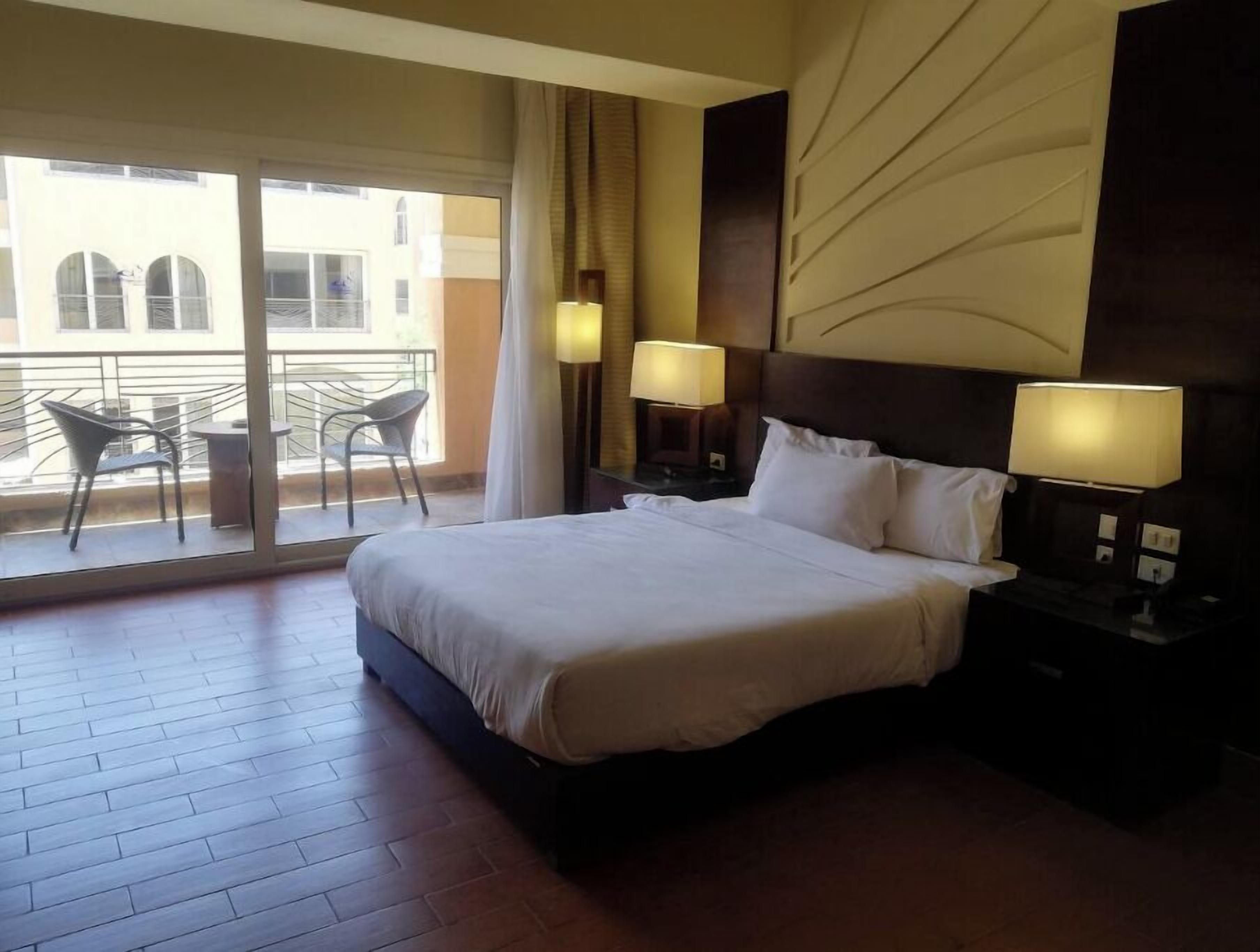 A hotel room with a large bed, two bedside tables with lamps, a balcony with chairs, and a view of buildings.