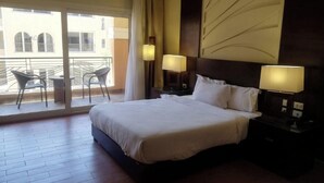 Habitación de hotel con una cama grande, dos mesitas de noche con lámparas, un balcón con sillas y vistas a edificios.
