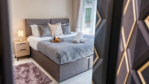 A modern bedroom with a large bed, a nightstand, a fluffy rug, and a window with curtains. - Maple House (Solihull)