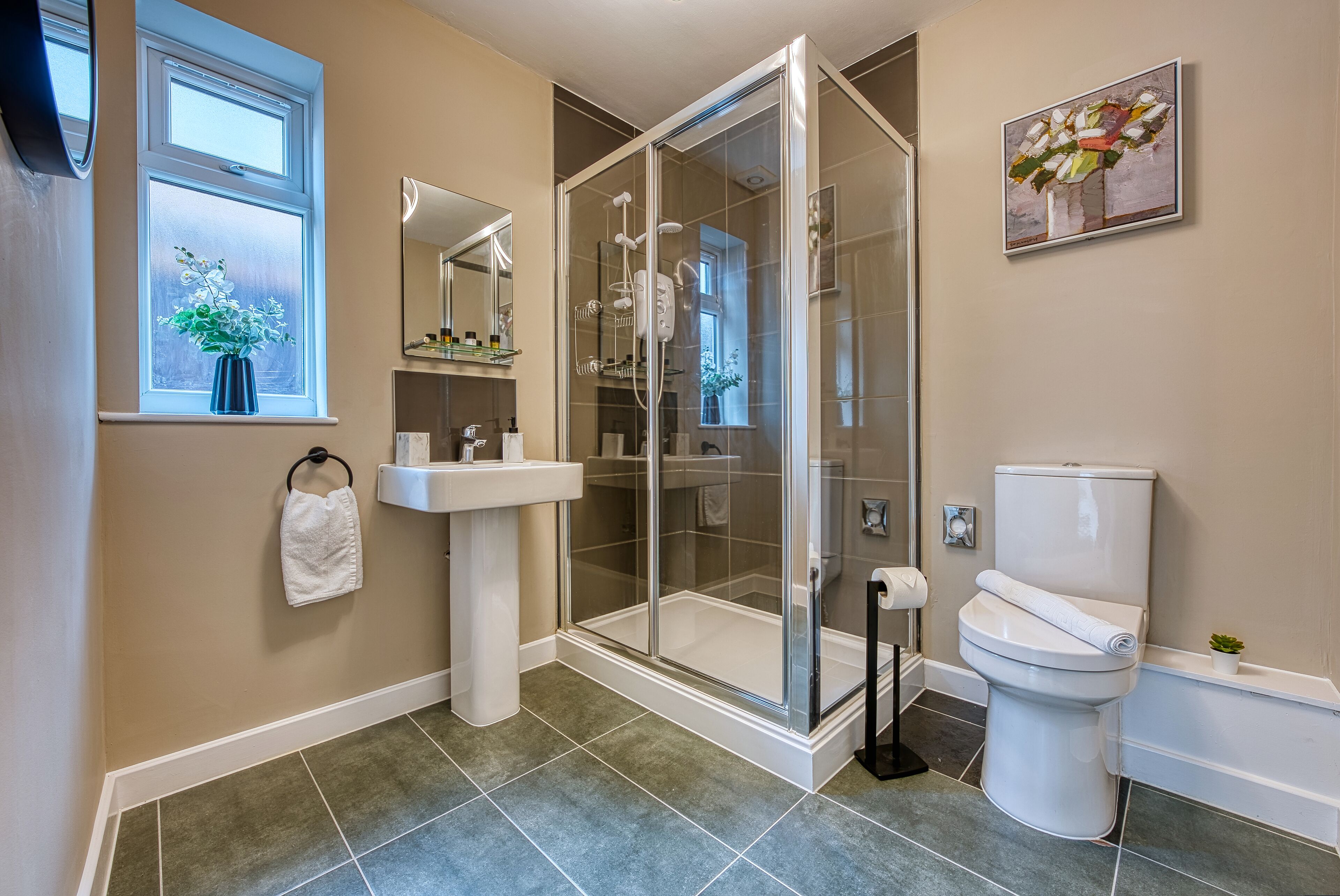 Une salle de bain avec une douche à parois de verre, un lavabo sur colonne, des toilettes et une fenêtre agrémentée d’une plante.