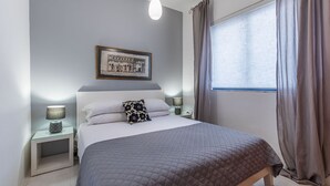 Ein ordentlich eingerichtetes Schlafzimmer mit einem Bett, Nachttischlampen, einem gerahmten Bild an der Wand und einem Fenster mit Vorhängen.