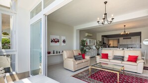 Ein modernes Wohnzimmer mit einem Glastisch, zwei Sofas mit roten Kissen, einem roten Teppich mit Muster und einer Küche im Hintergrund.