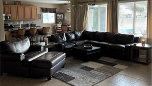 Een woonkamer met een modulaire bank, een poef en een salontafel. Er is een keuken met kasten en een eettafel met stoelen. Een plafondventilator is zichtbaar, en de kamer heeft grote ramen met gordijnen.