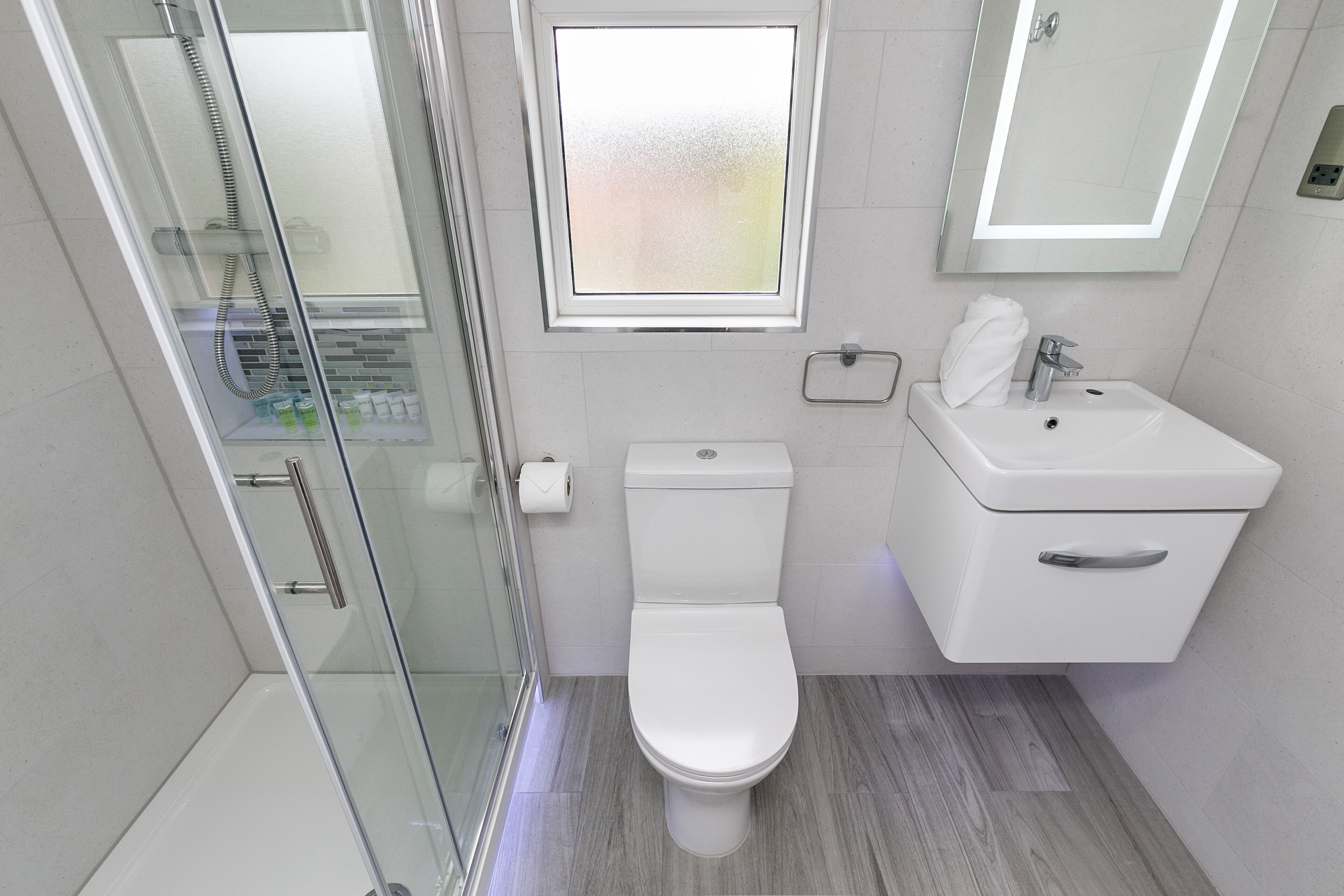 A modern bathroom with a glass shower enclosure, a toilet, a sink, and a window above the toilet.