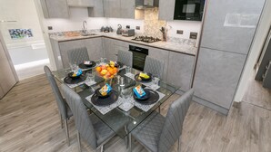 A modern kitchen with grey cabinetry, a glass dining table with chairs, and a central island with a fruit bowl. - Maple Lodge (Reading)