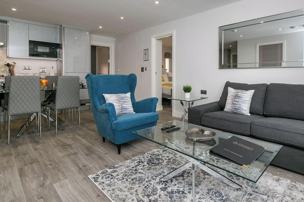 A modern living room with a grey sofa, a blue armchair, a glass coffee table, and a dining area with a glass table and chairs.