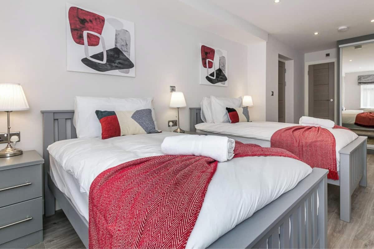 A hotel room with two beds, each with a red and white patterned blanket, white bedding, and a grey headboard. There are two bedside tables with lamps, a large mirror, and artwork on the walls.