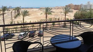 Un balcón con vistas a una playa de arena, palmeras y coches aparcados.