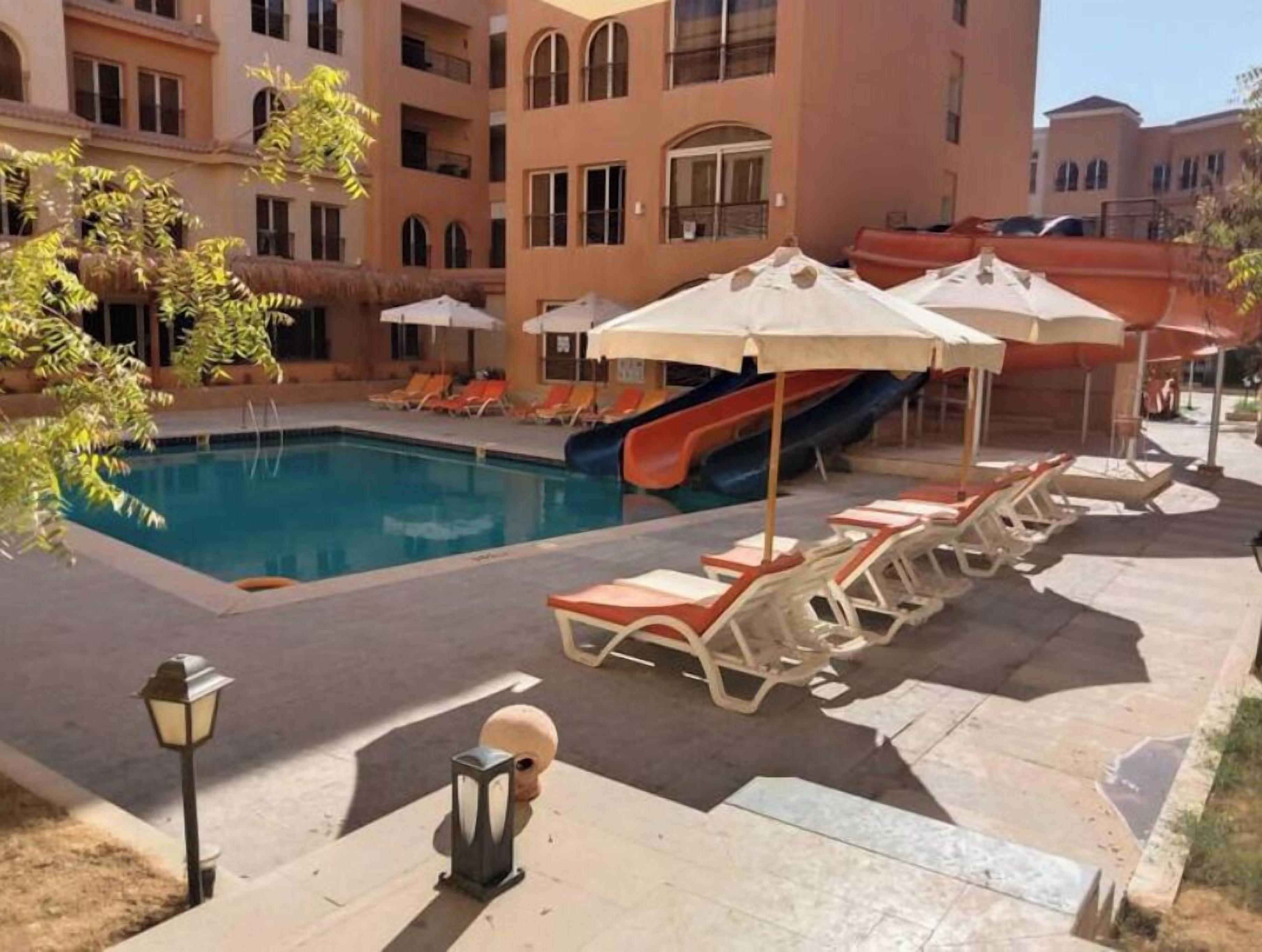 A swimming pool with lounge chairs and umbrellas, a slide, and a building with multiple balconies.