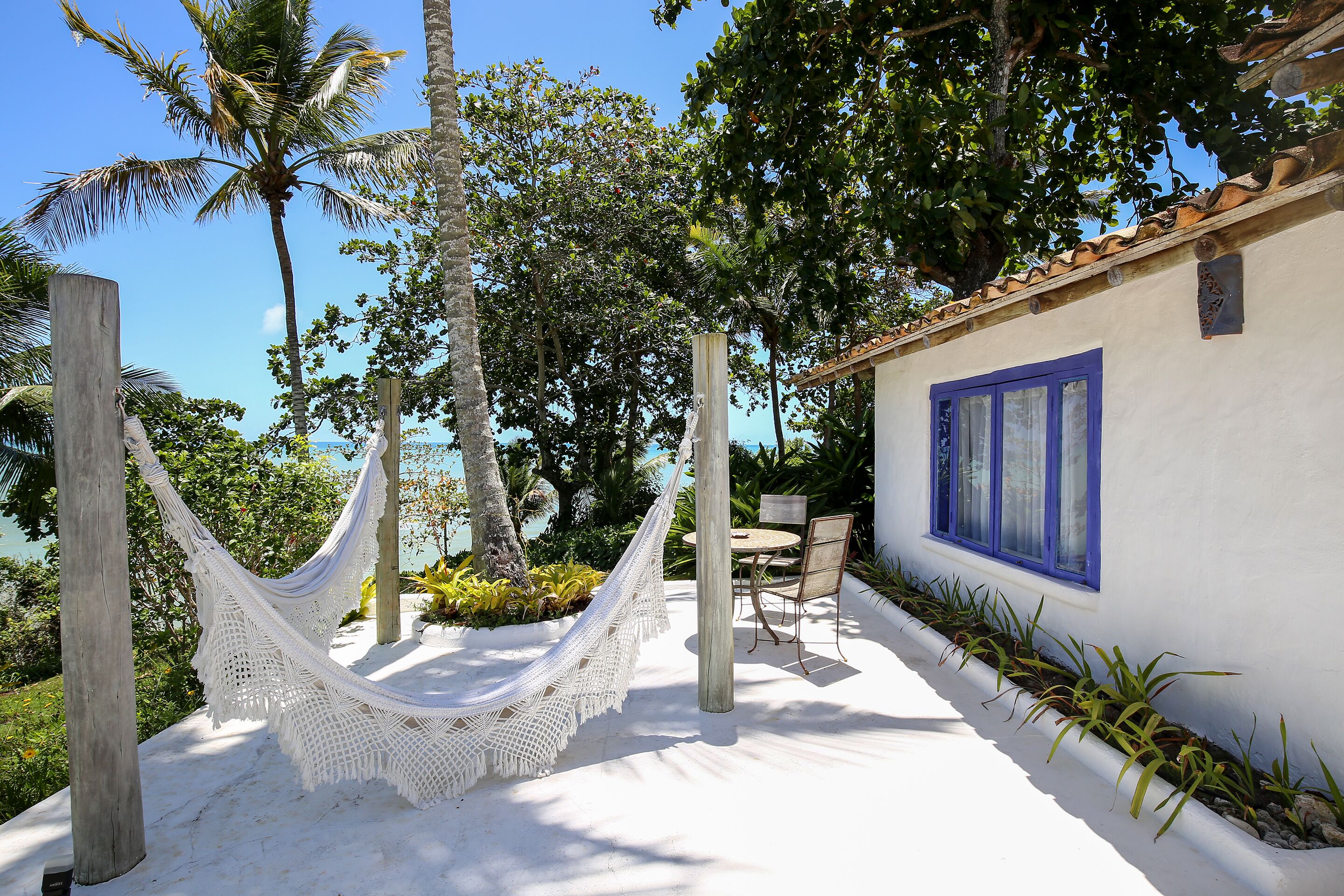 A hammock tied between wooden posts, a table and chairs, and a building with blue-framed windows.